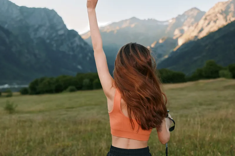 A cheerful woman stands outdoors in a scenic landscape, raising her hand in celebration while wearing an orange sports top. Distant mountains and an open meadow create a vibrant, inspirational