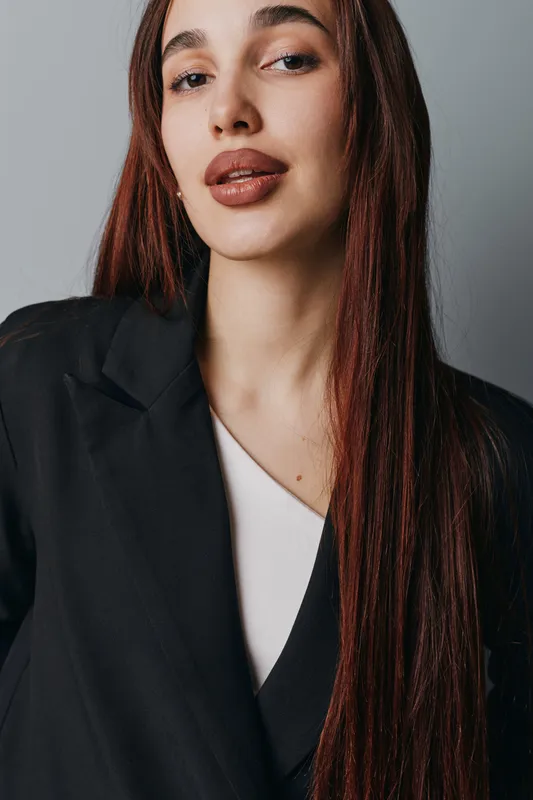 Woman with long straight hair wearing black blazer and white top posing against gray background with confident expression and natural makeup in studio portrait.