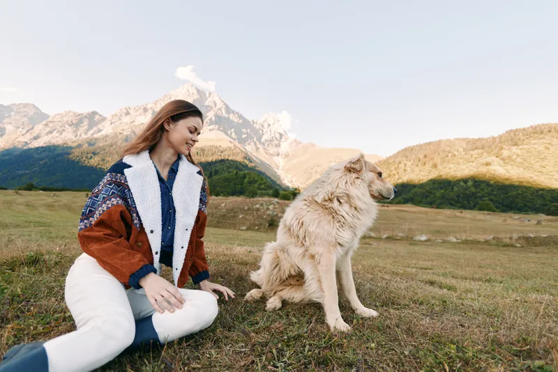 Woman and dog in mountains meadow outdoor scene, sitting and smiling as pet enjoys nature. Casual autumn clothing, relaxed pose, scenic landscape and soft natural light.