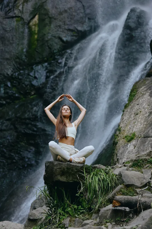young woman meditating near waterfall in white activewear, sitting cross-legged on rock, serene nature background, peaceful outdoor wellness moment