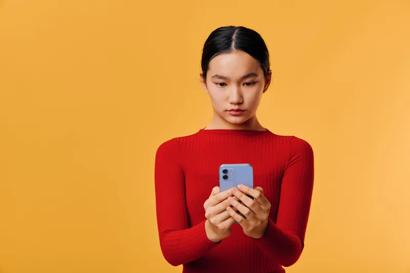 Young woman wearing red sweater looking at smartphone with concentration on orange background for technology, communication, social media and modern lifestyle concept.