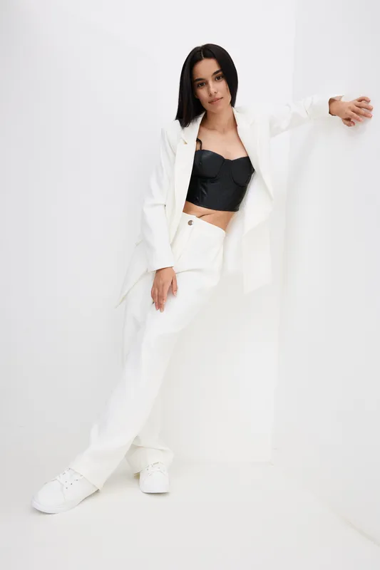 Young woman in stylish white suit and black crop top leaning on white wall, wearing sneakers, posing with relaxed confidence in minimalistic bright studio setting.