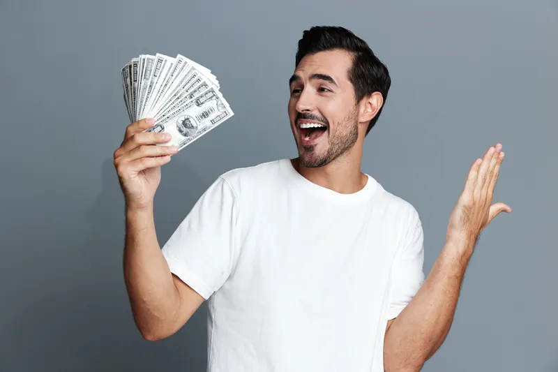 Excited smiling man holding cash money, feeling joyful and successful, dressed in white t-shirt, against grey background in studio portrait setting.