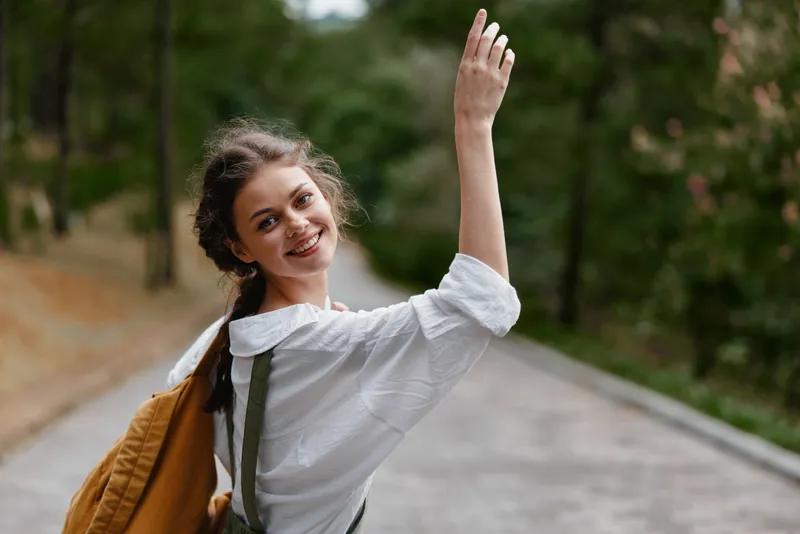 Young woman smiling and waving in a natural park setting, wearing a white shirt and backpack, exuding joy and warmth against a green forest backdrop