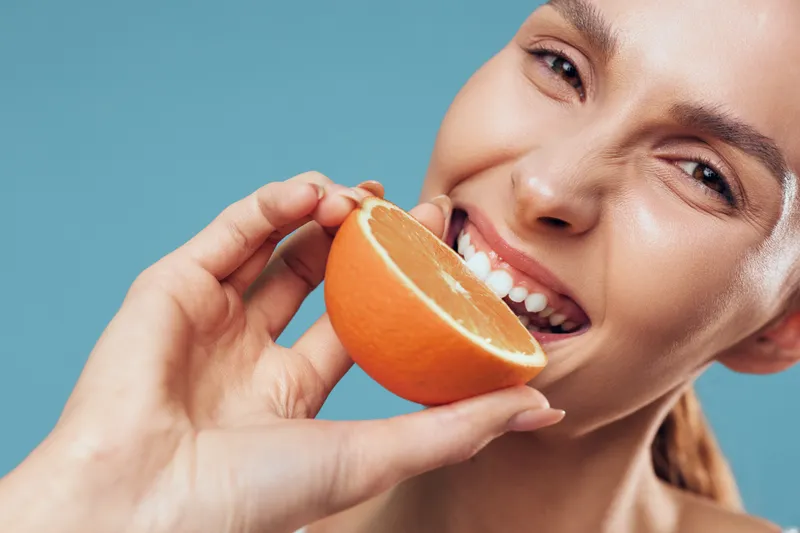 Woman eating orange in a closeup, smiling while holding citrus segments, fresh healthy vitamin nutrition concept on a plain blue background.