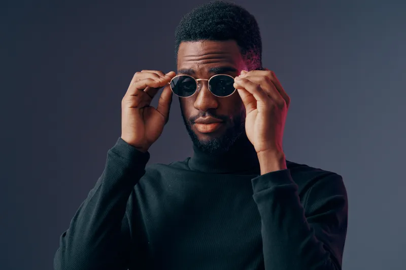 Stylish young African man in black turtleneck adjusting sunglasses, expressing confidence against a moody gray background with a modern vibe