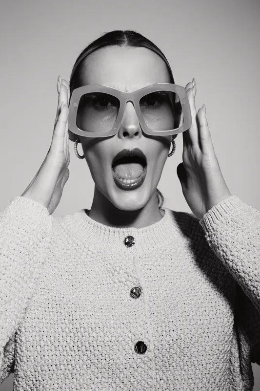 A surprised white woman with pale skin wearing oversized sunglasses and a knitted cardigan expresses shock with open mouth in a black and white studio portrait.