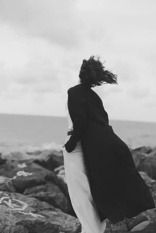 Wind swept silhouette of a figure in a long coat on a rocky shore by the ocean, dramatic movement of flowing fabric, moody landscape, solitude and outdoor fashion atmosphere.