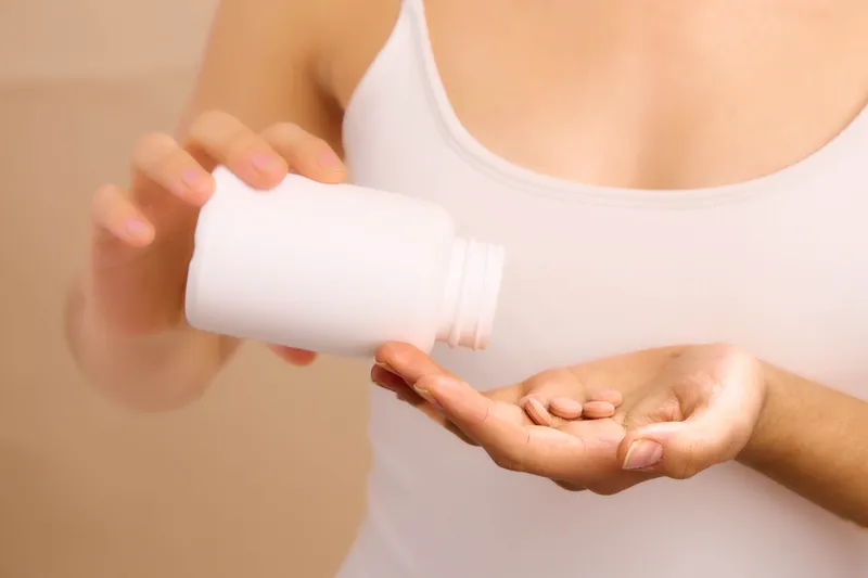 Asian woman wearing a simple tank top pours pills from a bottle into her hand, conveying a calm health care moment in a quiet home setting.