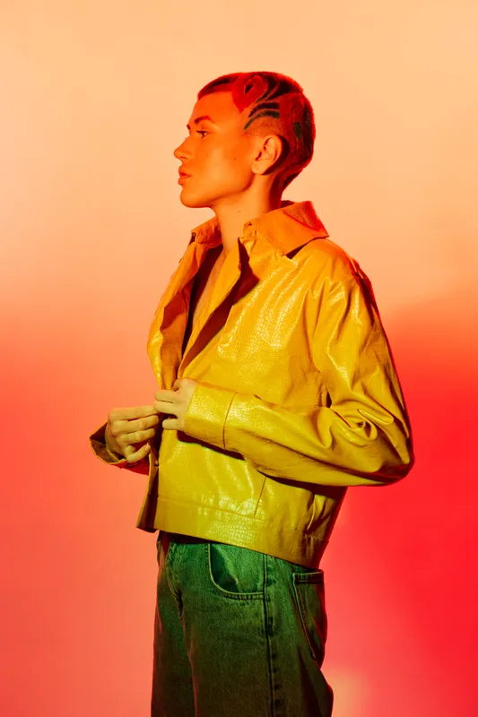 Young woman with short, artistic hairstyle in yellow jacket and denim jeans stands confidently against vibrant orange background, expressing modern style and creativity.