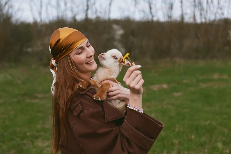 Cheerful woman outdoors, holding a goat kid, smiling at a flower, sunlit moment, relaxed posture, natural colors, grassy landscape, warm breeze, lifestyle bond