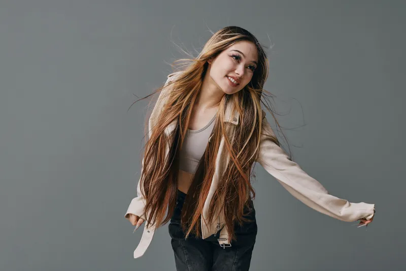 Young woman with long hair poses and smiles in casual outfit against grey background. Female model wearing light jacket and crop top in studio portrait with natural expression.