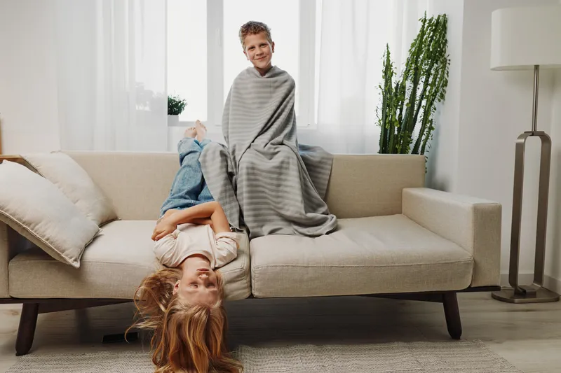 Two playful children spend time together on a beige sofa in bright living room, wrapped in a blanket and laughing, creating a warm and joyful family moment indoors.