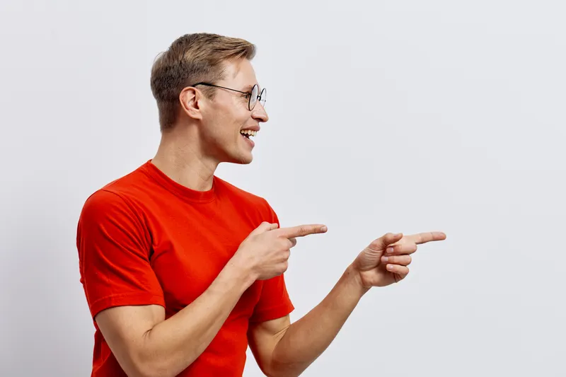 Smiling young caucasian man wearing red t-shirt and glasses points to the right with both hands, looking happy and cheerful isolated on plain white background. People lifestyle concept