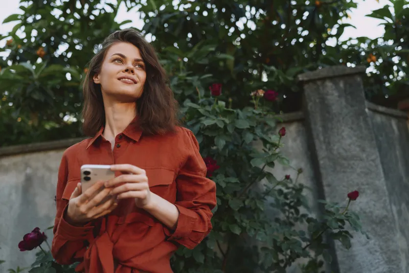 Woman lifestyle portrait of a relaxed young woman in a rust shirt holding a smartphone, standing in a garden with roses, casual mood and thoughtful gaze