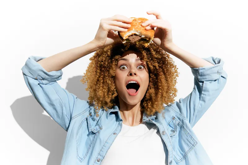 Взволнованная молодая женщина с вьющимися волосами счастливо держит вкусный бургер, выражая радость и энтузиазм