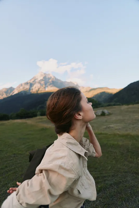 Young woman enjoys peaceful moment outdoors in nature with mountains in background wearing casual shirt and backpack during sunny day.