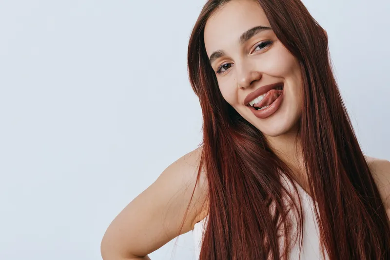 Young woman with long brown hair smiling playfully and sticking out her tongue against a plain light background. Casual portrait with natural expression and relaxed pose.