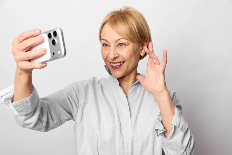 Smiling senior woman takes a smartphone selfie, waving. Friendly smile and communication, technology portrait on a white studio background, casual shirt, indoors.