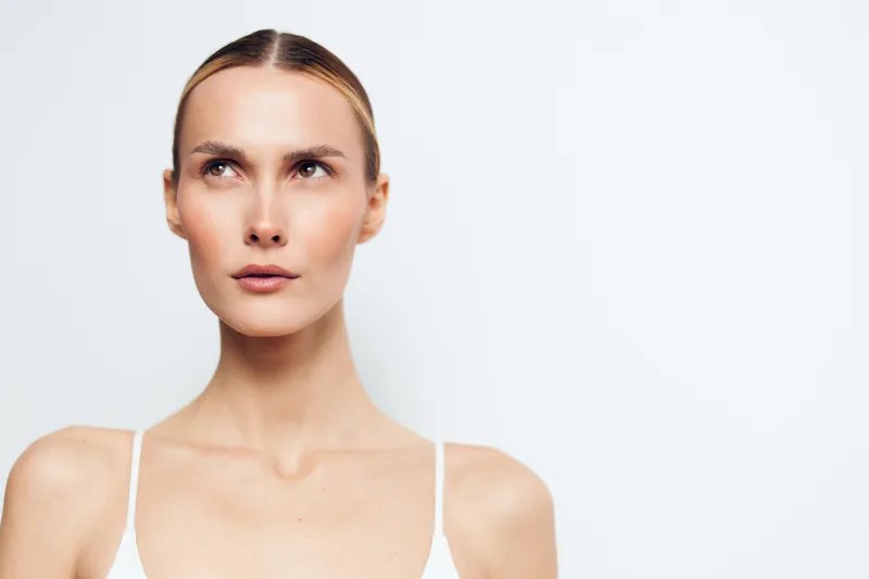 Portrait of a serious woman with smooth skin and slicked back hair looking up. Minimalist white background emphasizes natural beauty and clean skin concept in soft lighting.
