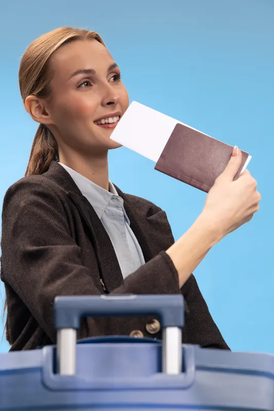 Young smiling woman holds passport and boarding pass near blue suitcase. Travel concept with happy tourist preparing for trip, vacation, and adventure on light blue background.