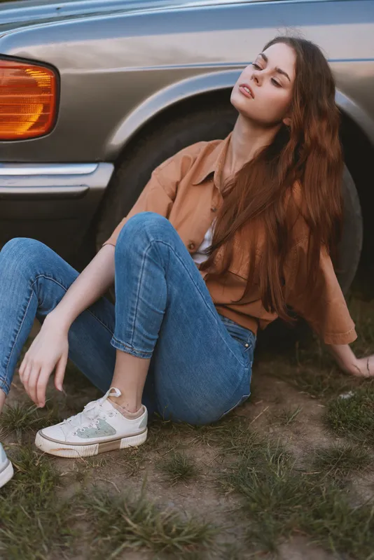 Young woman in casual outfit with long hair relaxing by a vintage car in a natural setting. Bright colors and serene mood capture a carefree lifestyle.