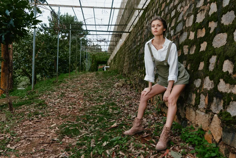 Young woman in a green outfit posing outdoors against a stone wall, evoking nature and confidence, with a calm atmosphere in a garden setting