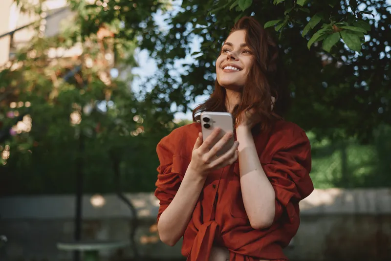 Happy woman outdoors lifestyle with smartphone in park, casual red blouse, smiling portrait, candid moment in sunlight