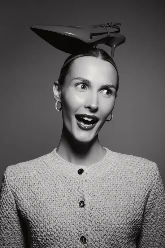 Caucasian woman with light skin wears high heel shoe on head, smiling with earrings in black and white studio portrait. Concept of fashion, creativity, and fun.