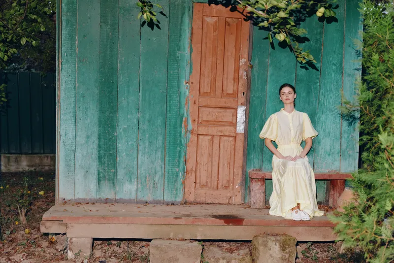 Woman in a pale yellow dress stands on a wooden porch in front of a weathered teal wall, serene outdoor scene with natural surroundings