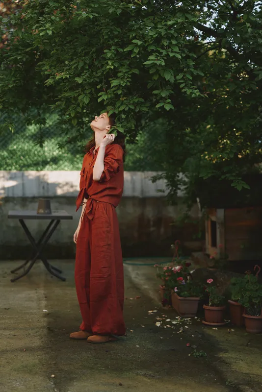 Woman in red dress stands under a lush tree in a garden, enjoying a calm lifestyle moment, sunlit leaves, casual outdoor portrait, serene everyday scene