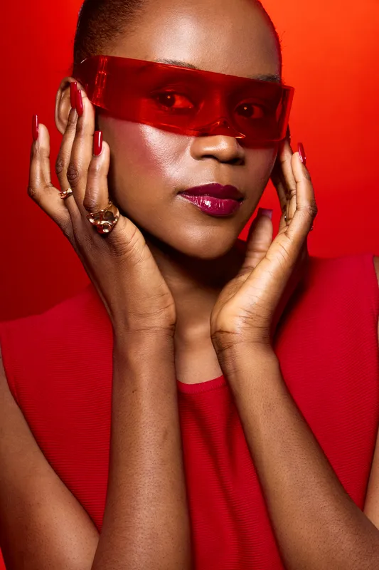 Portrait of confident black woman with dark skin wearing red dress and translucent red glasses. Fashion makeup with glossy lips and red nails. Concept of elegance, style, and beauty.