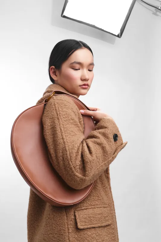 asian woman wearing a brown wool coat poses with a round leather bag slung over her shoulder in a bright studio, exuding calm confidence and poised elegance.
