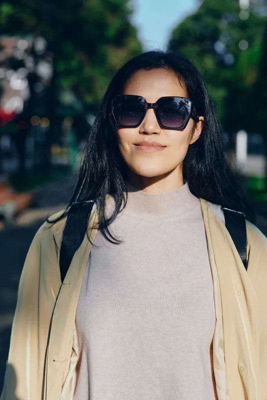 Stylish woman wearing oversized sunglasses and light sweater with a beige coat, backpack over shoulders, outdoors on a bright day, casual street style and confident mood in an urban setting
