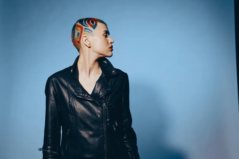 Woman with colorful shaved hair and black leather jacket looking sideways against blue background, expressing bold style and modern attitude.