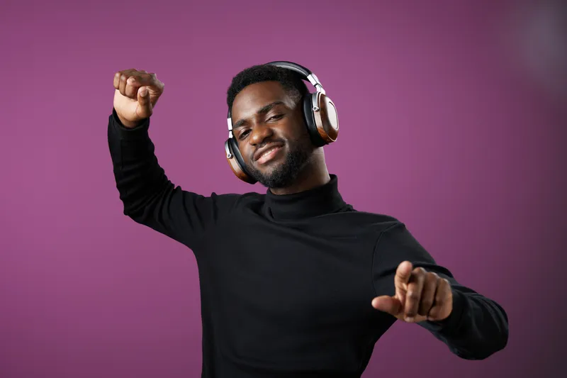 Young Black man enjoying music with headphones, dancing joyfully against a purple background in a stylish outfit, emphasizing happiness and relaxation