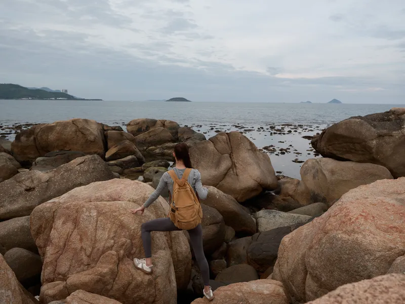 Young female hiker exploring rocky coastline, wearing outdoor attire, with a neutral expression, in serene coastal landscape, concept of adventure and exploration
