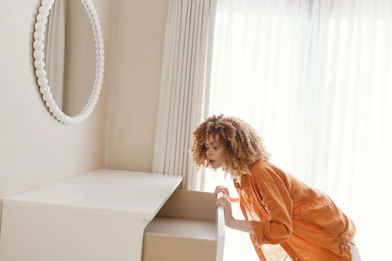 На изображении любопытная женщина с вьющимися волосами в оранжевой рубашке открывает ящик в светлой минималистичной комнате