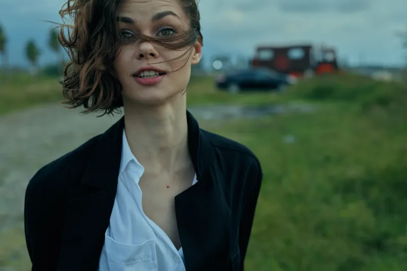 Woman outdoors with windblown hair, blazer and white shirt, standing on a grassy field with a distant blurred vehicle, expressive mood and moody light.