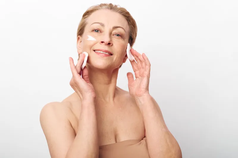 Middle aged woman applies face cream with cotton pads on cheeks smiling gently. Skincare routine, self care, beauty, and natural wellness on white background in bright soft lighting.