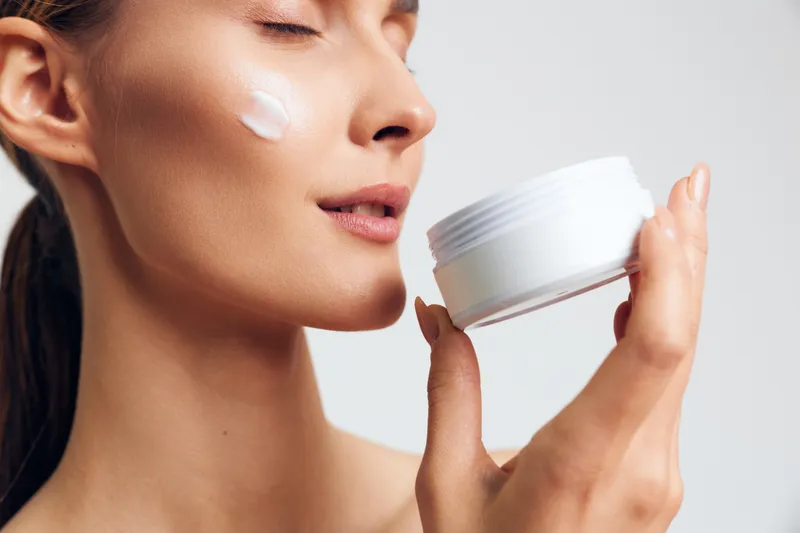 Closeup of woman with smooth skin holding white cream jar, applying moisturizer on cheek, natural beauty, skincare routine, soft lighting, neutral background.