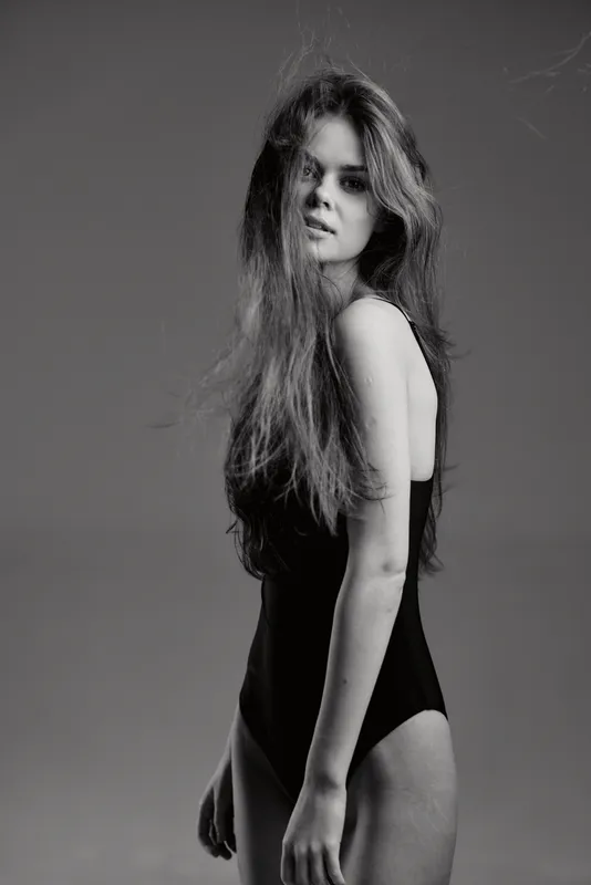 Black and white portrait of a serious woman with long hair wearing a black bodysuit against a plain background.