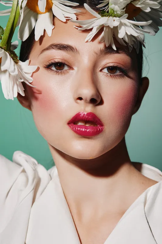 Close-up portrait of woman with bright pink lips and natural makeup wearing a daisy flower crown. Fashion beauty concept illustrating creativity and spring freshness on green background.