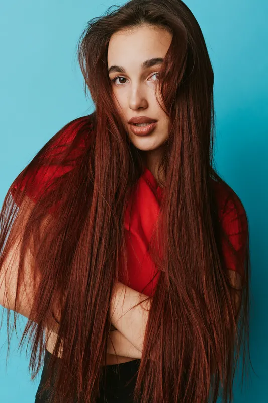 Woman with long straight hair and natural makeup wearing red shirt posing with crossed arms against blue background. Portrait of confident female model with casual style and expressive look.
