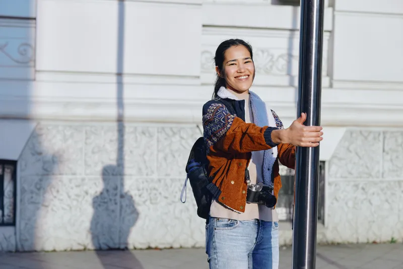 Smiling woman stands outdoors in an urban street, holding a pole, wearing a backpack, jacket and scarf, with sunlight on a bright day, conveying casual travel, youth energy and everyday exploration