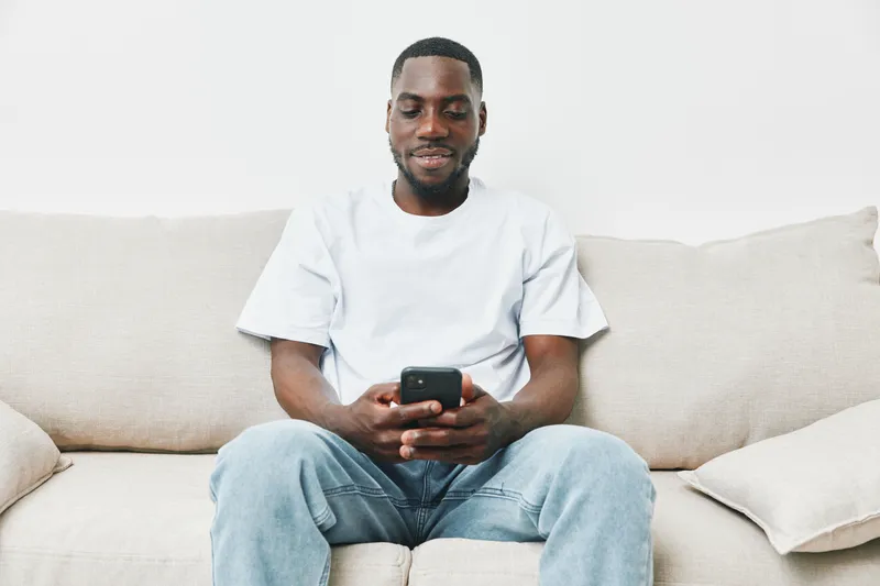 Young Black man sitting on a beige couch, smiling while using a smartphone, showcasing casual fashion and relaxed emotions in a modern living space