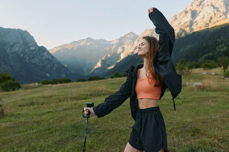 A fit young woman enjoying an outdoor hike stands in a mountainous meadow, raising her arm in triumph while gripping a trekking pole, radiating energy, confidence, and freedom.