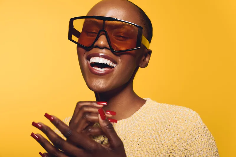 Happy African woman with dark skin and short hair wears large sunglasses and a yellow sweater. She smiles brightly against a vibrant yellow background. Concept of joy and fashion.