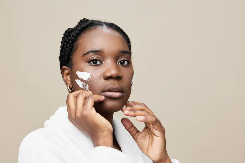 Young Black woman applying skincare cream on her face, showcasing beauty routine in a neutral toned setting