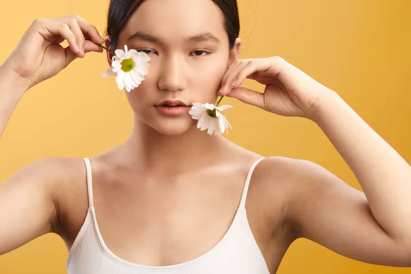 asian woman with white daisies held near her face stands against a bright yellow backdrop, soft lighting highlights smooth skin, natural makeup, and a calm, playful expression.